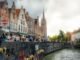 Picturesque scene of Bruges, showcasing historic buildings, a canal, and bustling crowds on a sunny day.