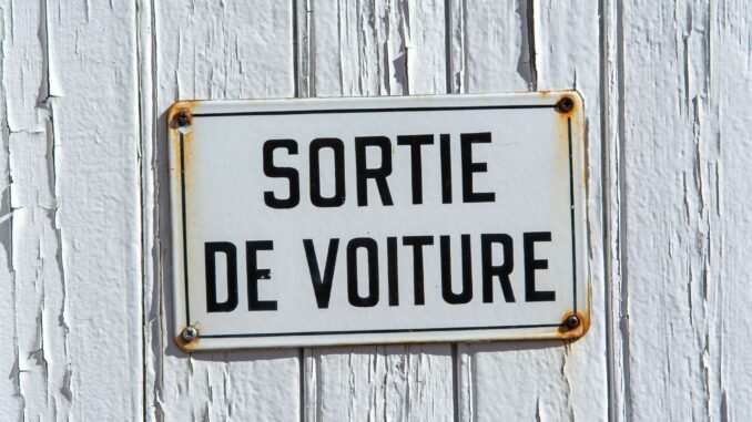 Close-up of a rustic French sign reading 'Sortie de Voiture' on a weathered white wooden wall.