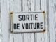 Close-up of a rustic French sign reading 'Sortie de Voiture' on a weathered white wooden wall.